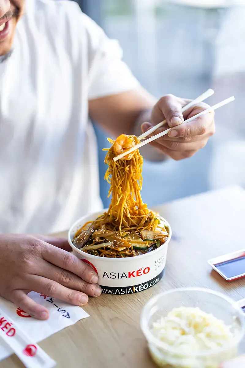 Une personne utilise des baguettes pour manger des nouilles dans un bol étiqueté « ASIA KEO ».