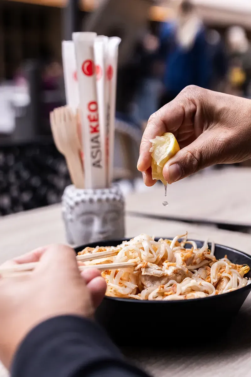 Une personne presse un citron au-dessus d'un bol de nouilles, de baguettes et d'un porte-baguettes en forme de tête de Bouddha.