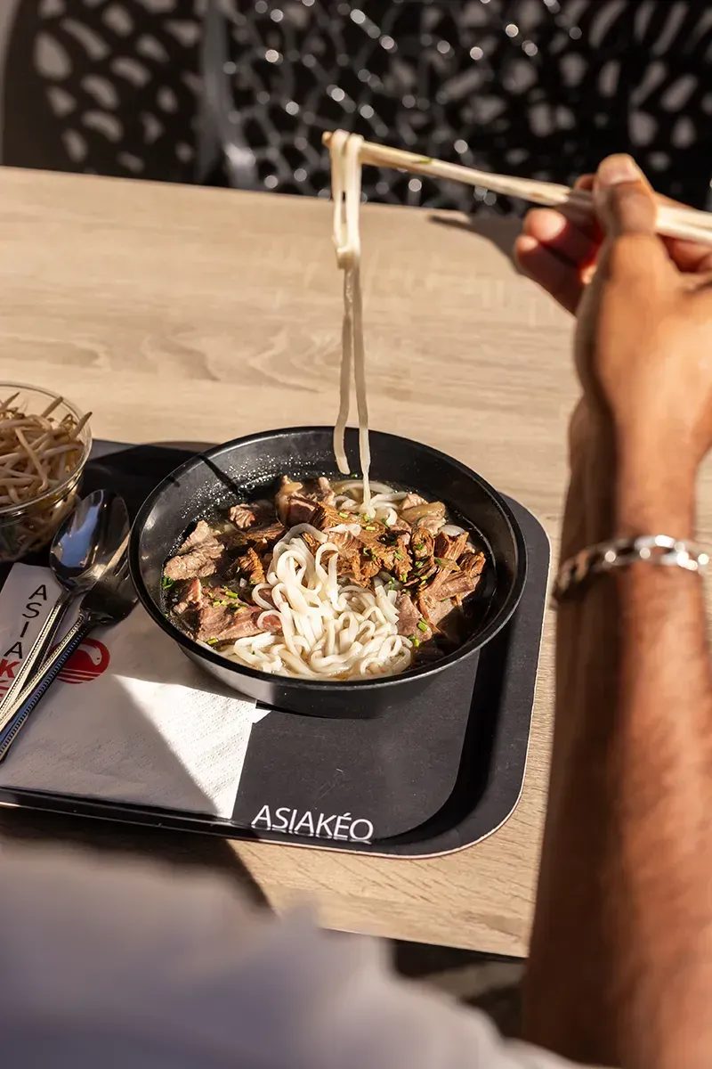 Une personne mange des ramen avec des baguettes, en extérieur. Elle prélève des nouilles d'un bol posé sur un plateau.