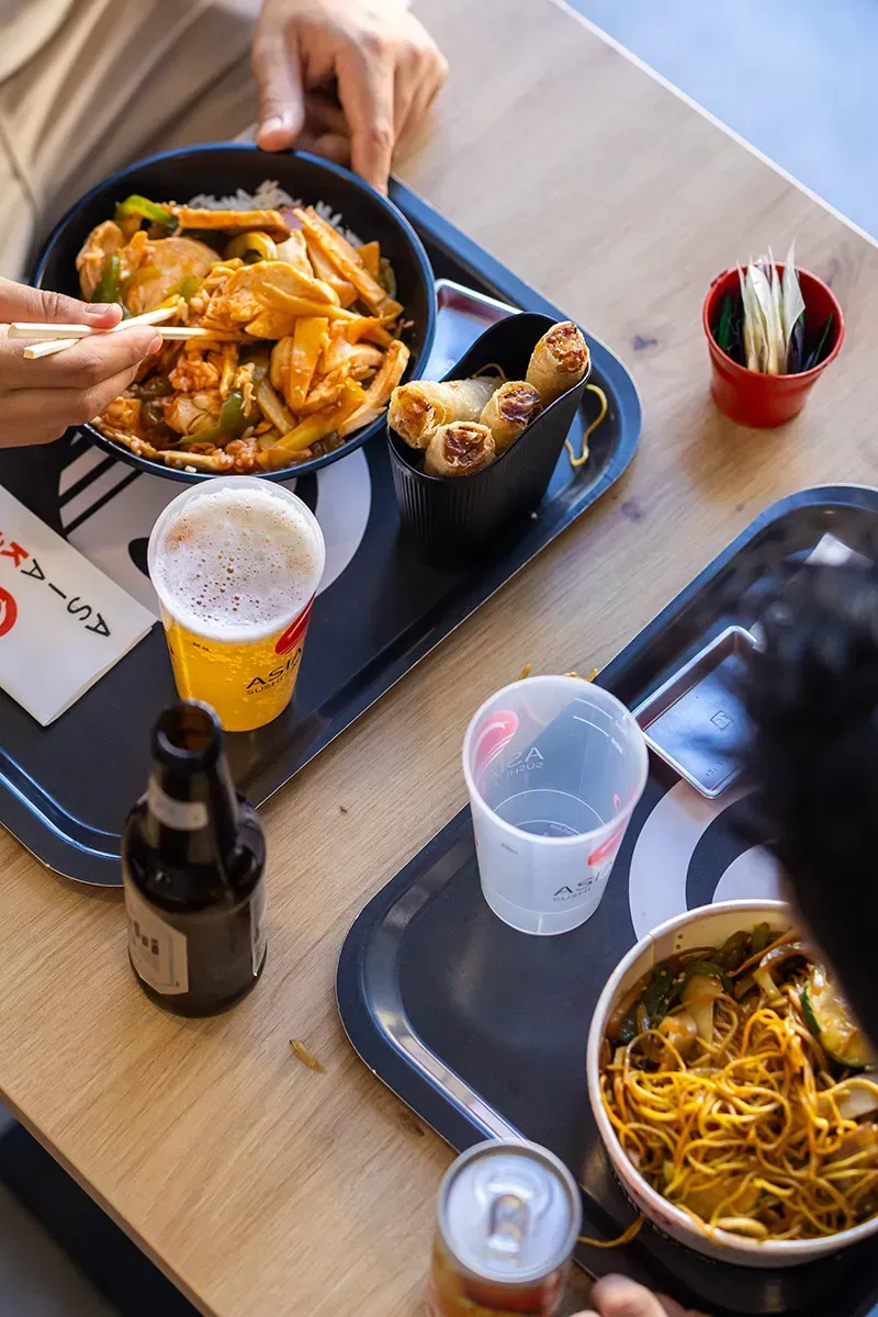 Deux plateaux de nourriture asiatique sont disposés sur une table en bois. Une personne mange dans un bol à l'aide de baguettes. On aperçoit de la bière et d'autres boissons.