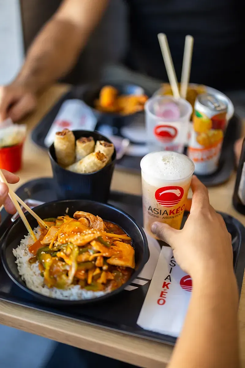 Plateau de nourriture asiatique : bol de nourriture avec baguettes, bière, rouleaux de printemps, boisson, personne tenant une bière.