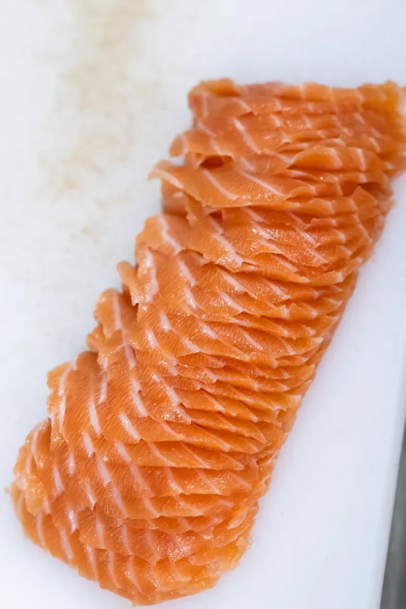Du saumon cru finement tranché sur une planche à découper blanche, mettant en valeur sa couleur orangée et sa texture.