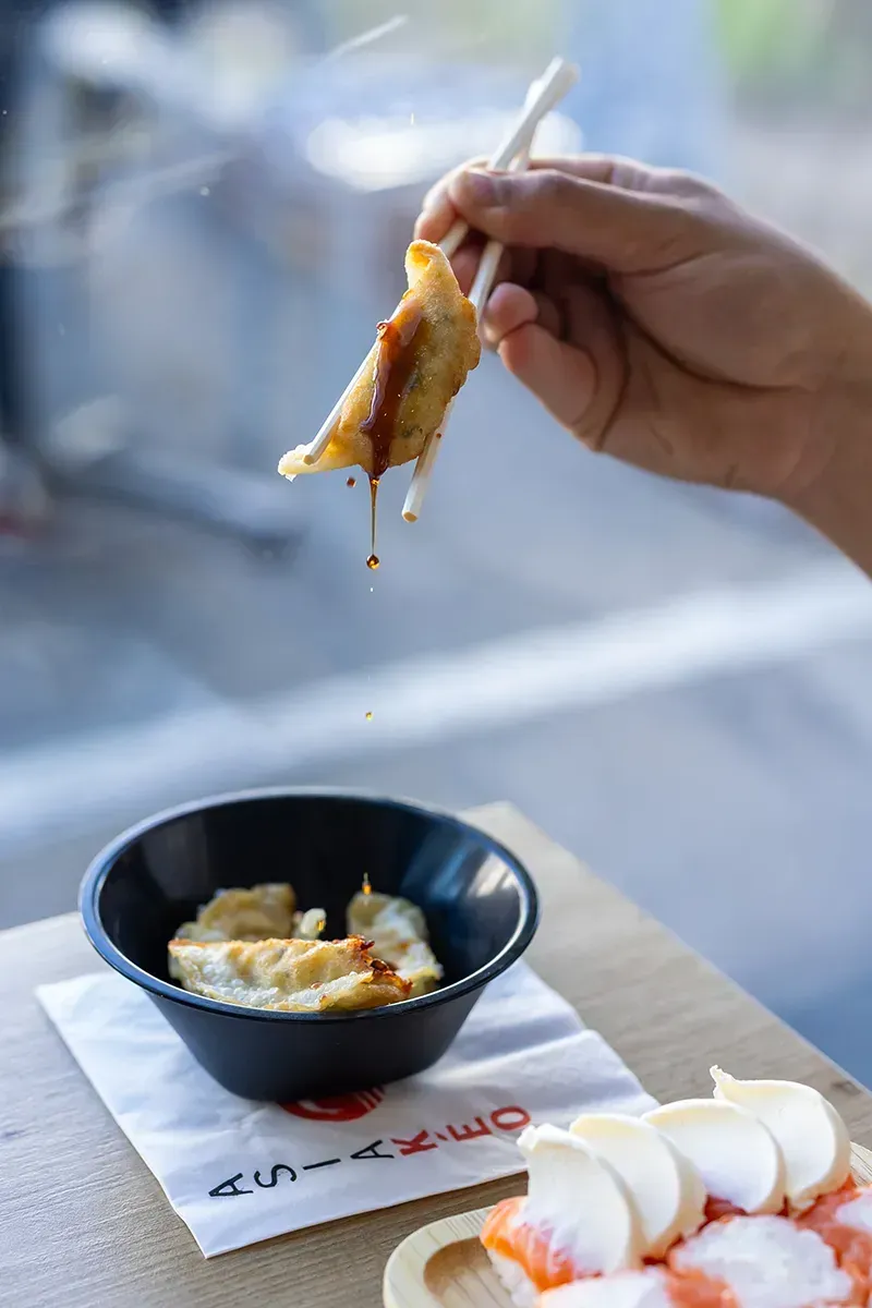 Une main utilise des baguettes pour tremper un ravioli dans la sauce, avec un bol de raviolis et d'autres aliments sur une table en bois.