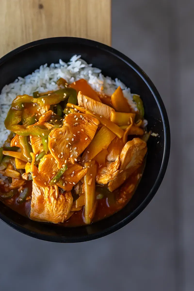 Bol de riz blanc garni de légumes orange et verts, et de poulet en sauce foncée.