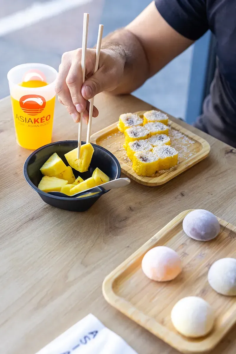 Une personne mange des morceaux d'ananas avec des baguettes, servis dans un bol avec des mochis et une boisson sur une table.