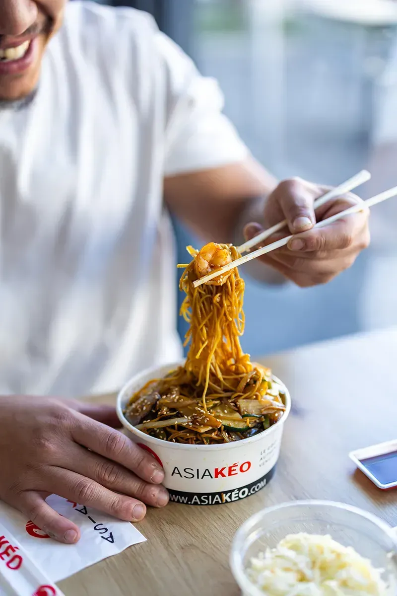 Une personne mange des nouilles asiatiques dans un bol, à l'aide de baguettes. L'inscription sur le bol est « ASIA KEO ».