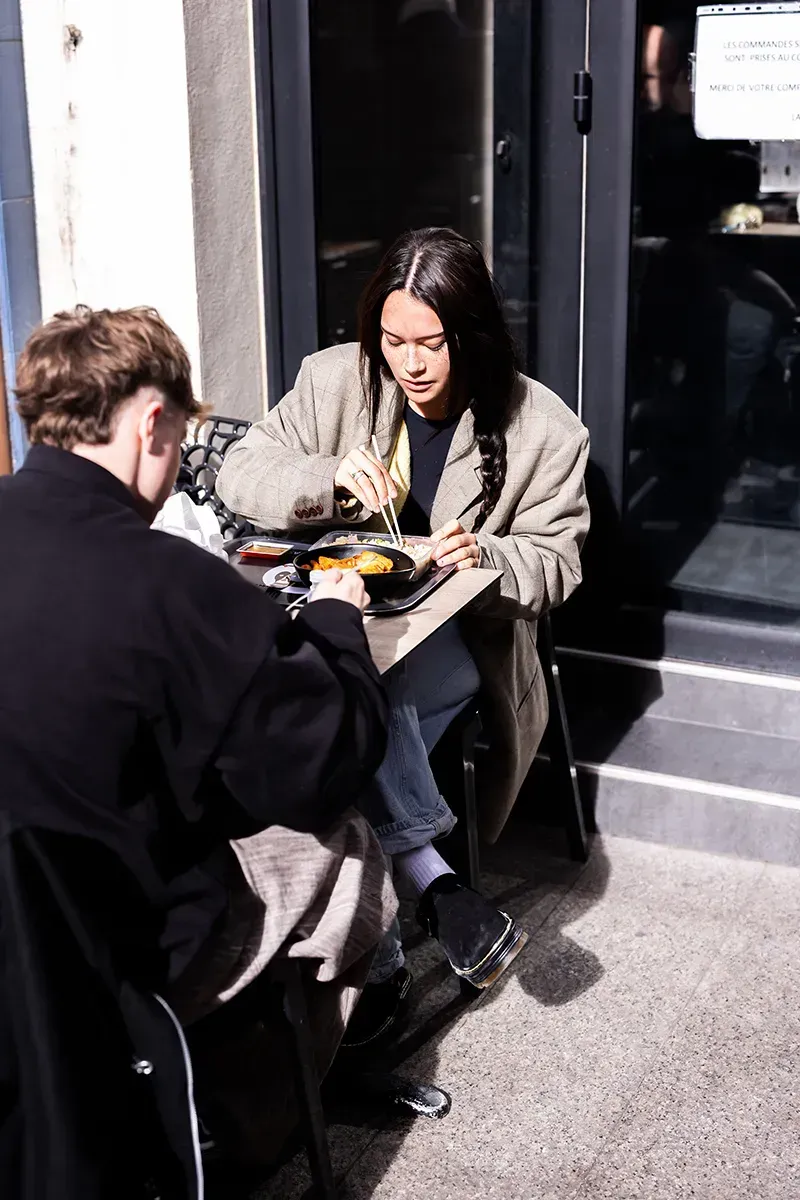 Deux personnes déjeunent à une table en terrasse. L'une porte un blazer et mange dans un récipient. L'autre, vêtue de noir, est en grande partie cachée.