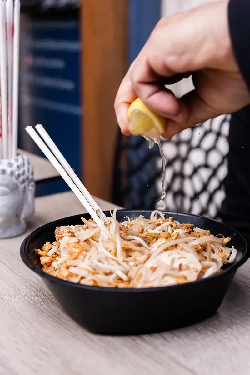 Une personne presse du jus de citron sur un bol de nouilles à l'aide de baguettes.