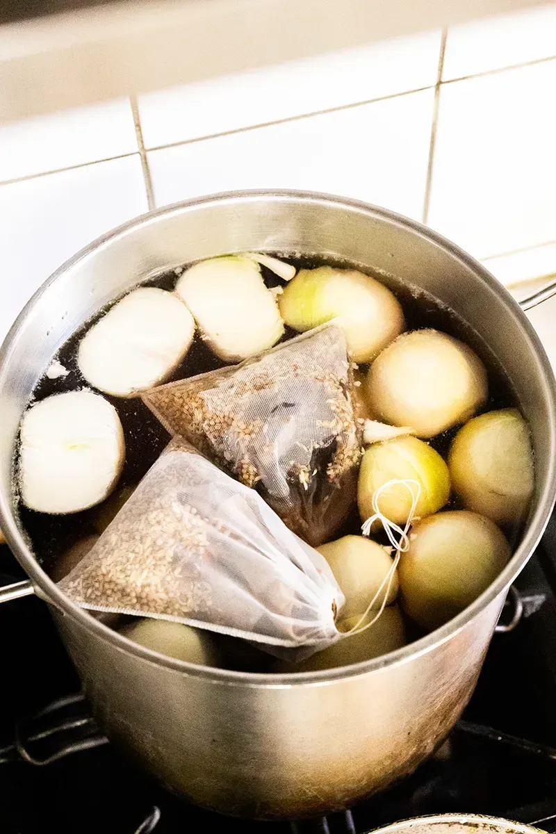 Une casserole de liquide bouillant avec des oignons et des sachets d'épices sur la cuisinière.