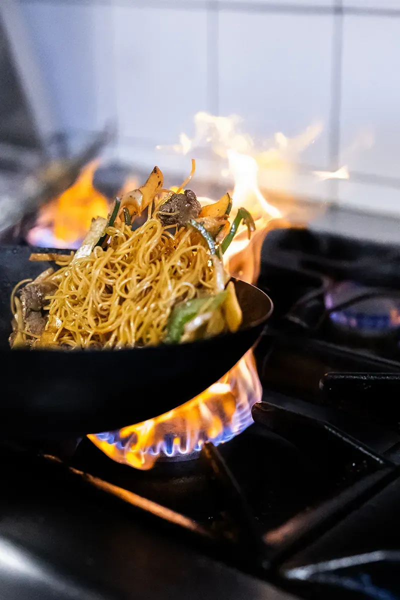 Wok contenant des nouilles et des légumes cuisant sur les flammes d'une cuisinière à gaz.