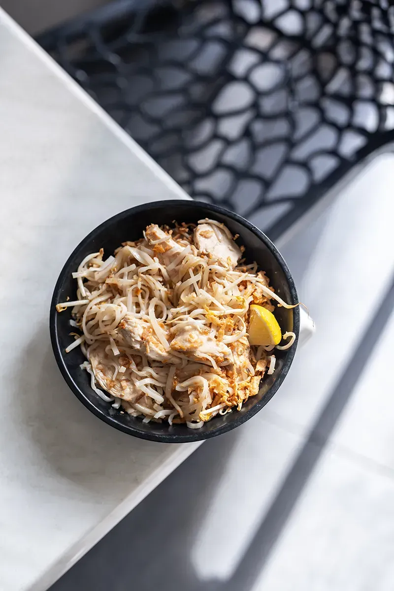 Pad thaï servi dans un bol noir avec un quartier de citron sur une table blanche.