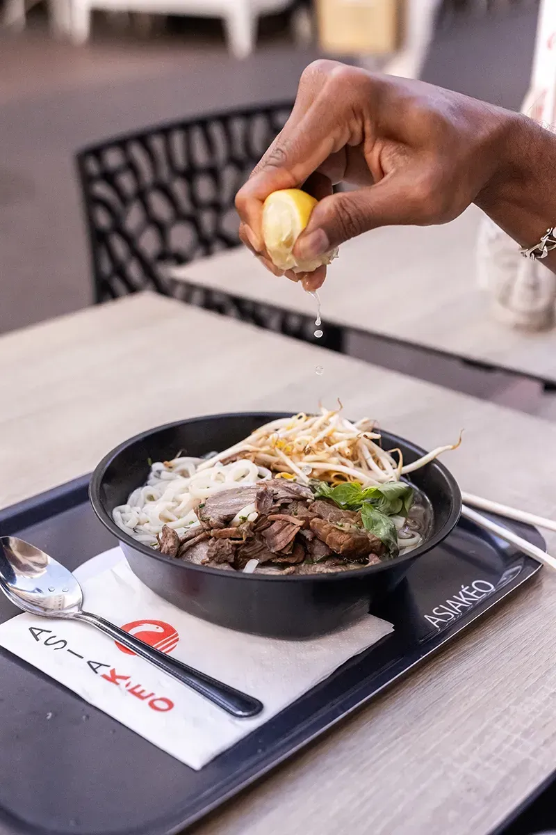 Une personne presse un citron au-dessus d'un bol de nouilles. La table est dressée avec des baguettes et une cuillère.