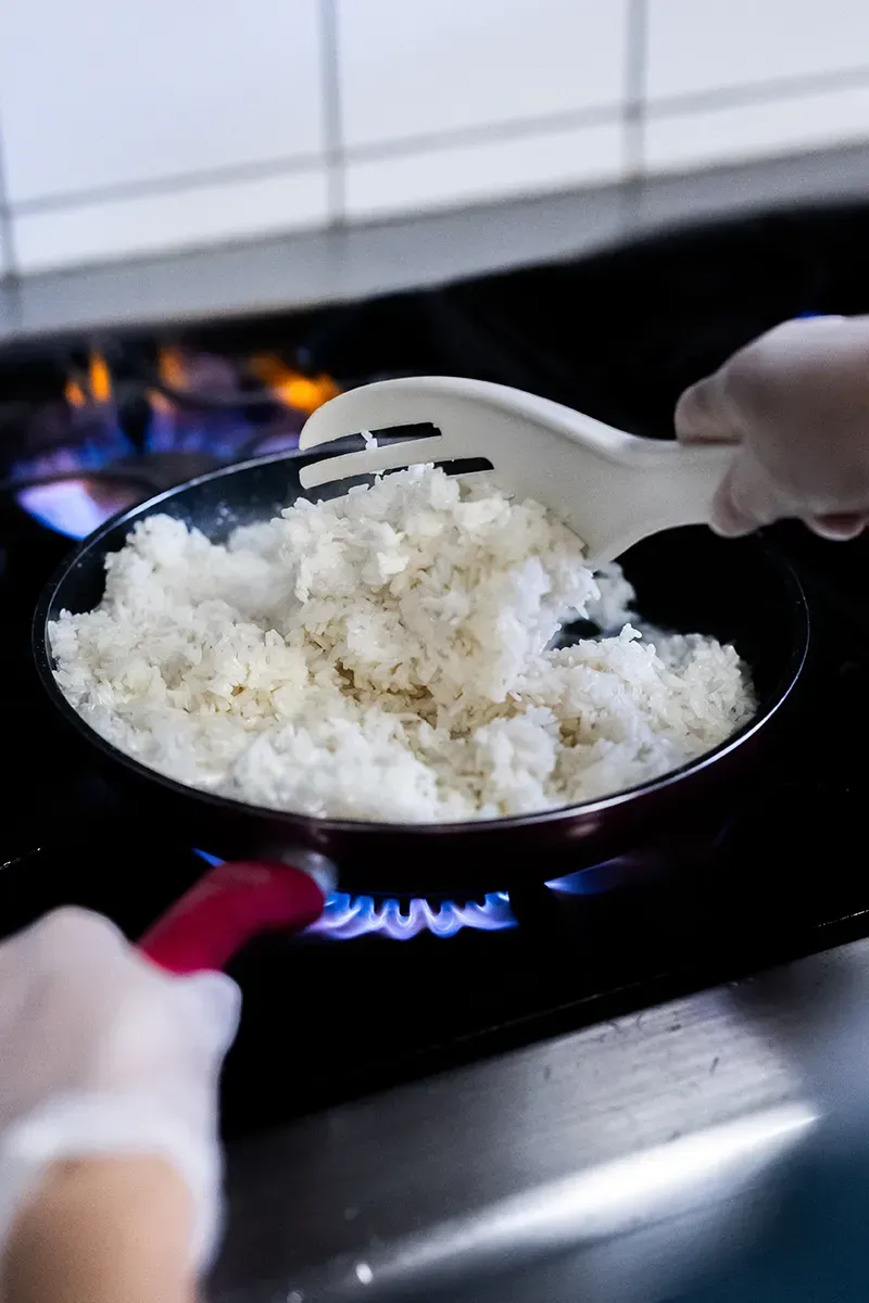 Une personne portant des gants remue du riz blanc dans une casserole sur une cuisinière à gaz.