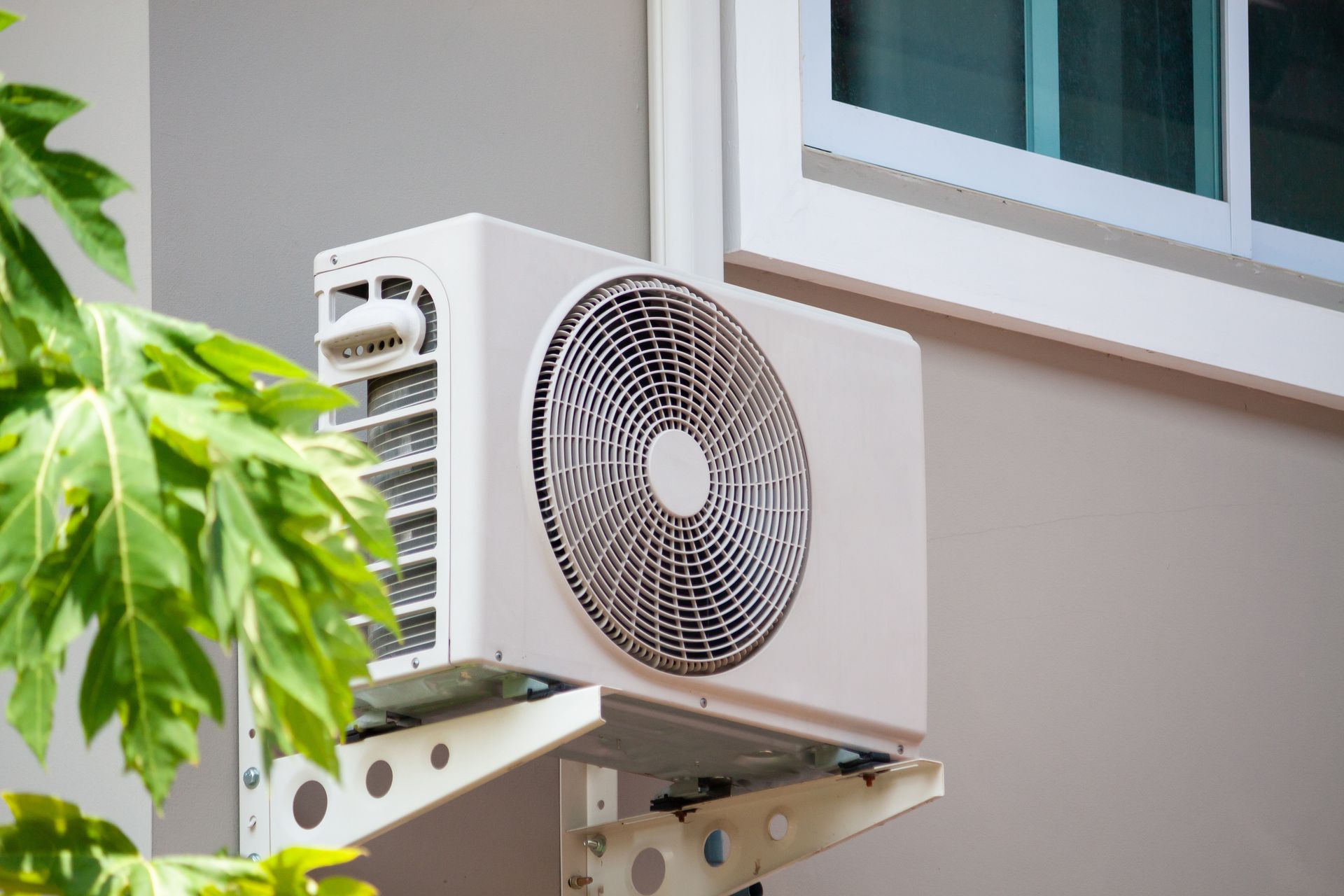 White air conditioning unit mounted on a gray wall, next to a window and green leaves.