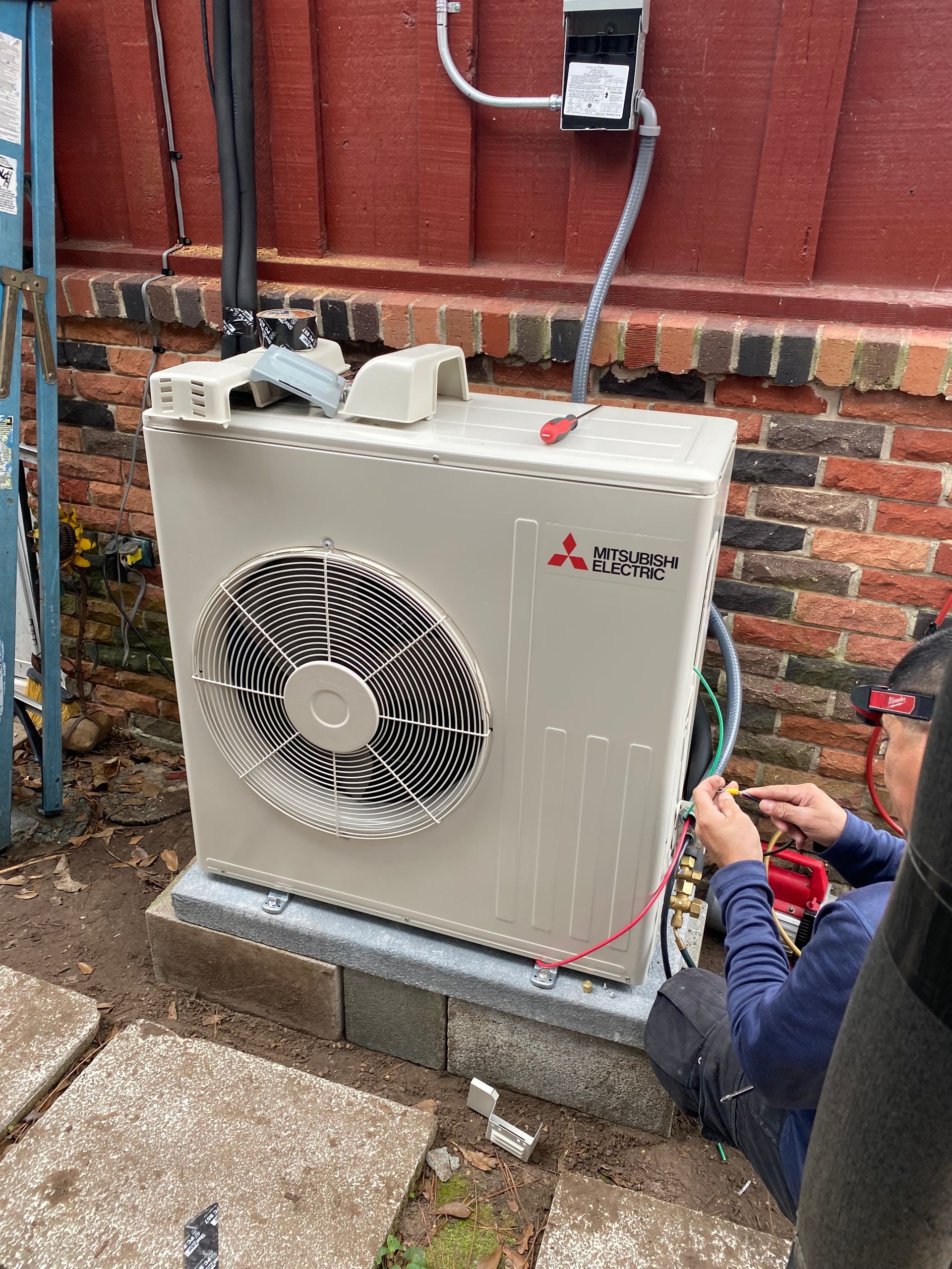 HVAC technician working on a Mitsubishi outdoor unit, mounted on a concrete pad near a brick wall.