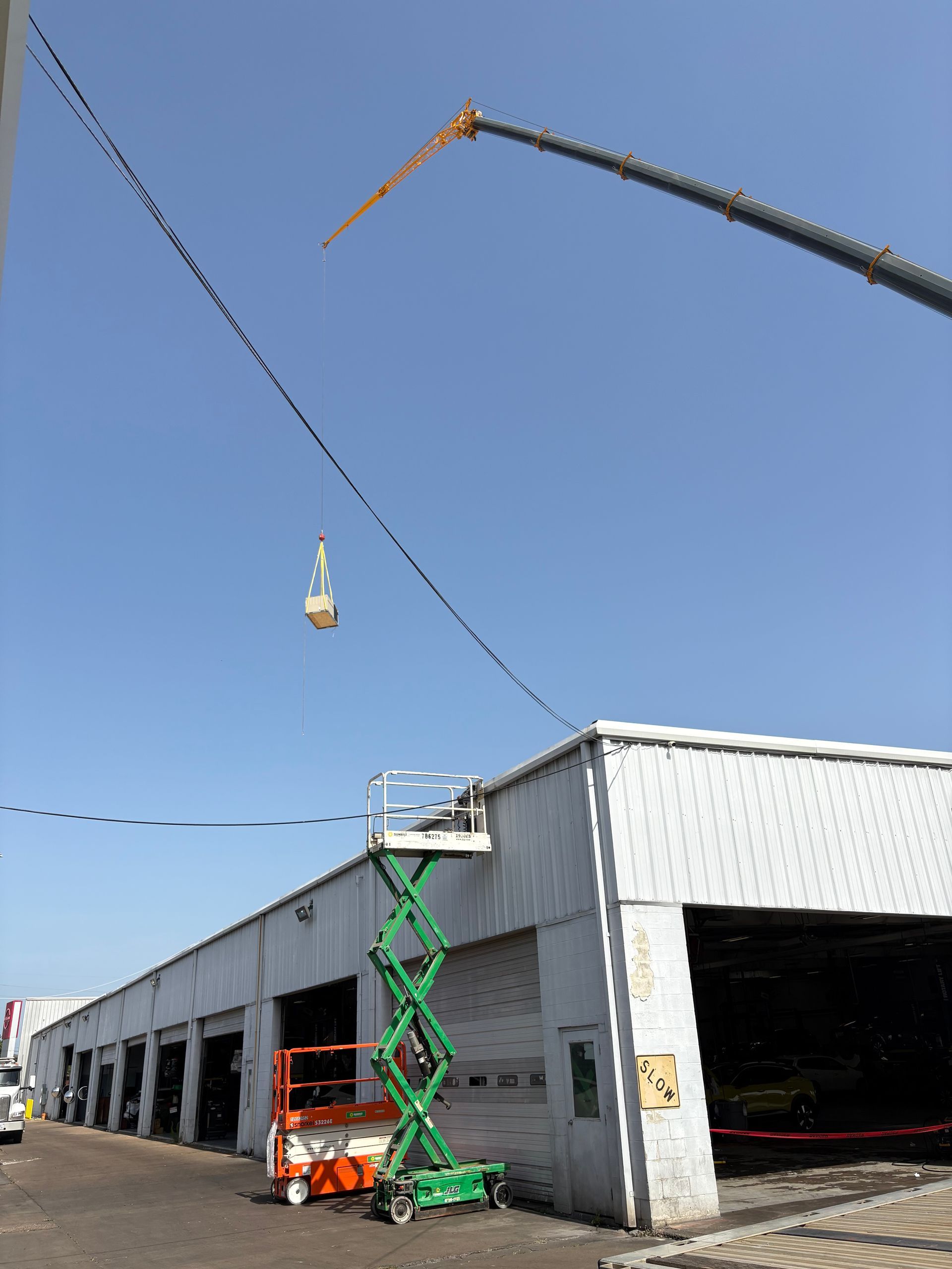 A crane lifting an object near a white warehouse with a green scissor lift.
