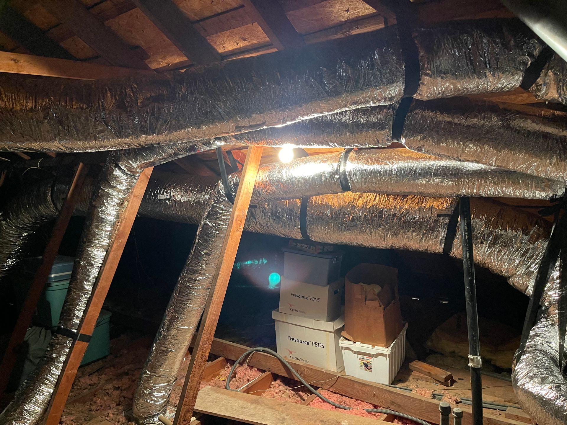 Attic interior with insulated ductwork, wooden beams, boxes, and a light.