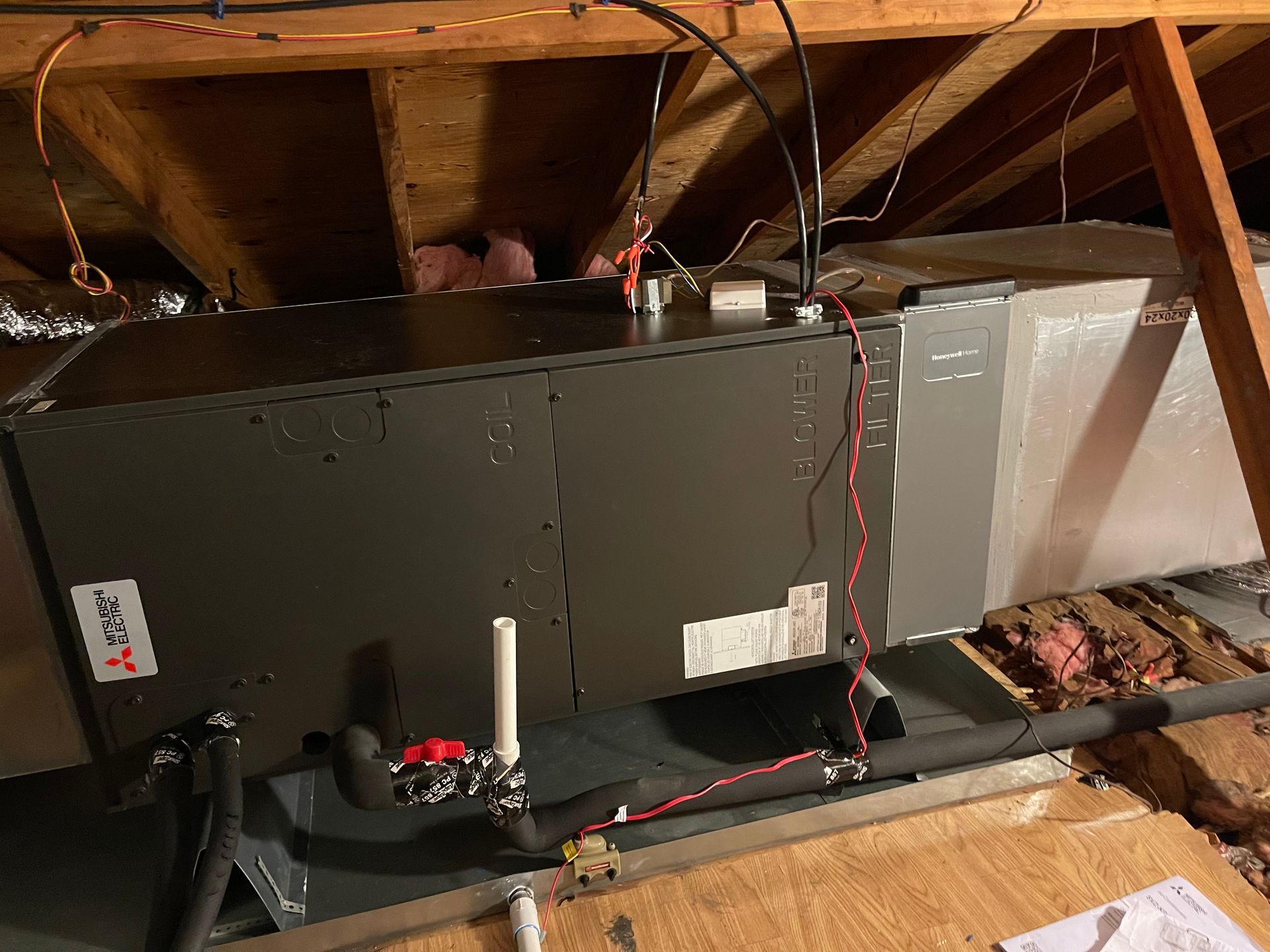 HVAC unit in an attic with ductwork, electrical wiring, and plumbing visible. Wooden beams and insulation surround it.