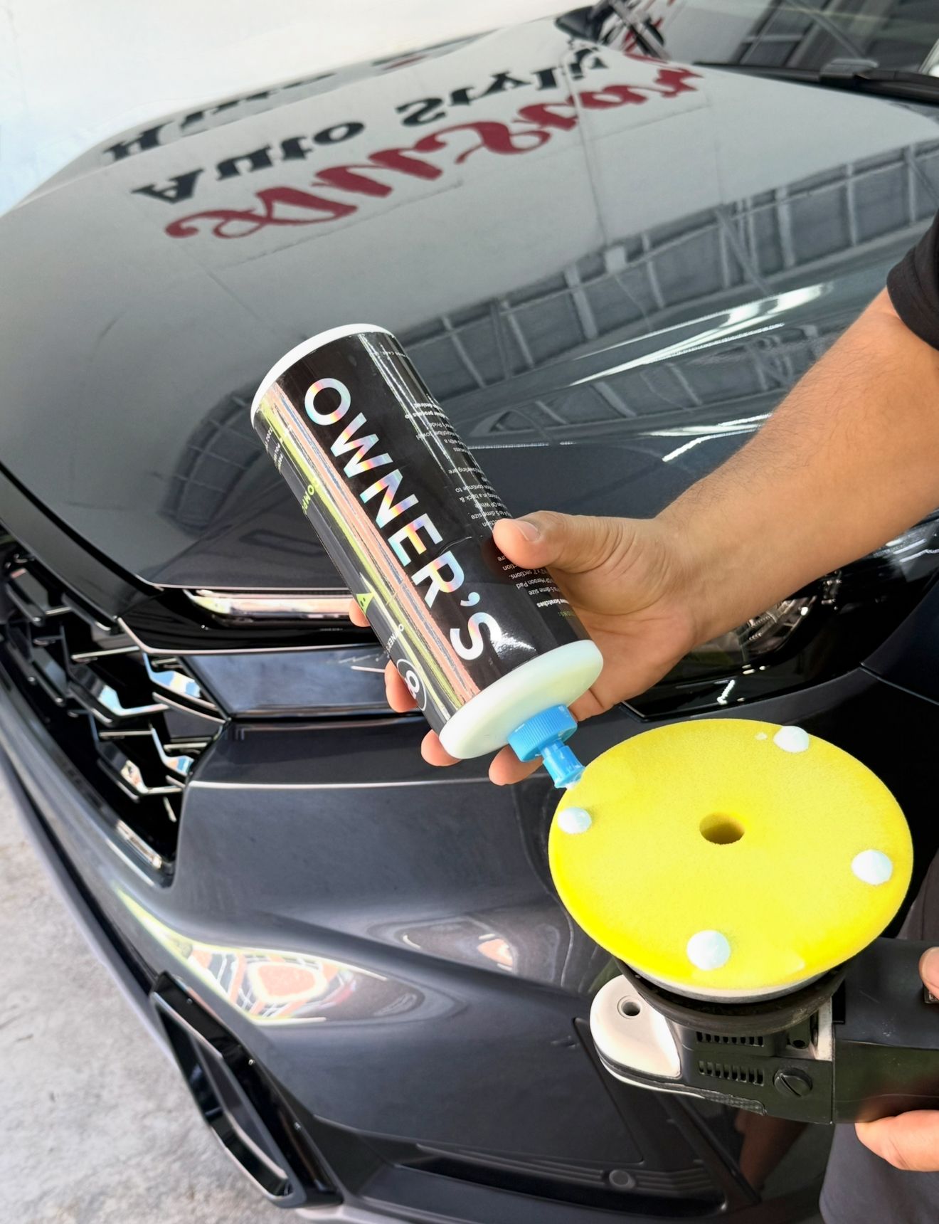 A person applies car polish from a bottle to a yellow buffer on a dark gray car.