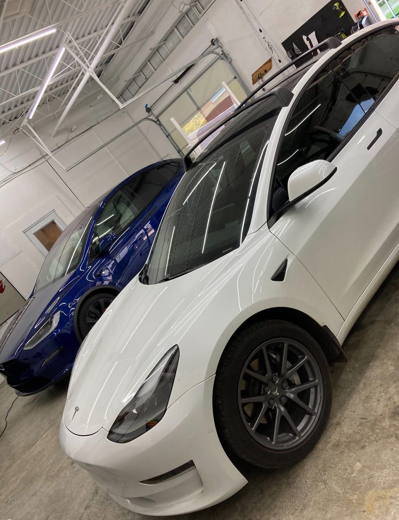 Two Tesla cars, white and blue, parked inside a garage with overhead lights.