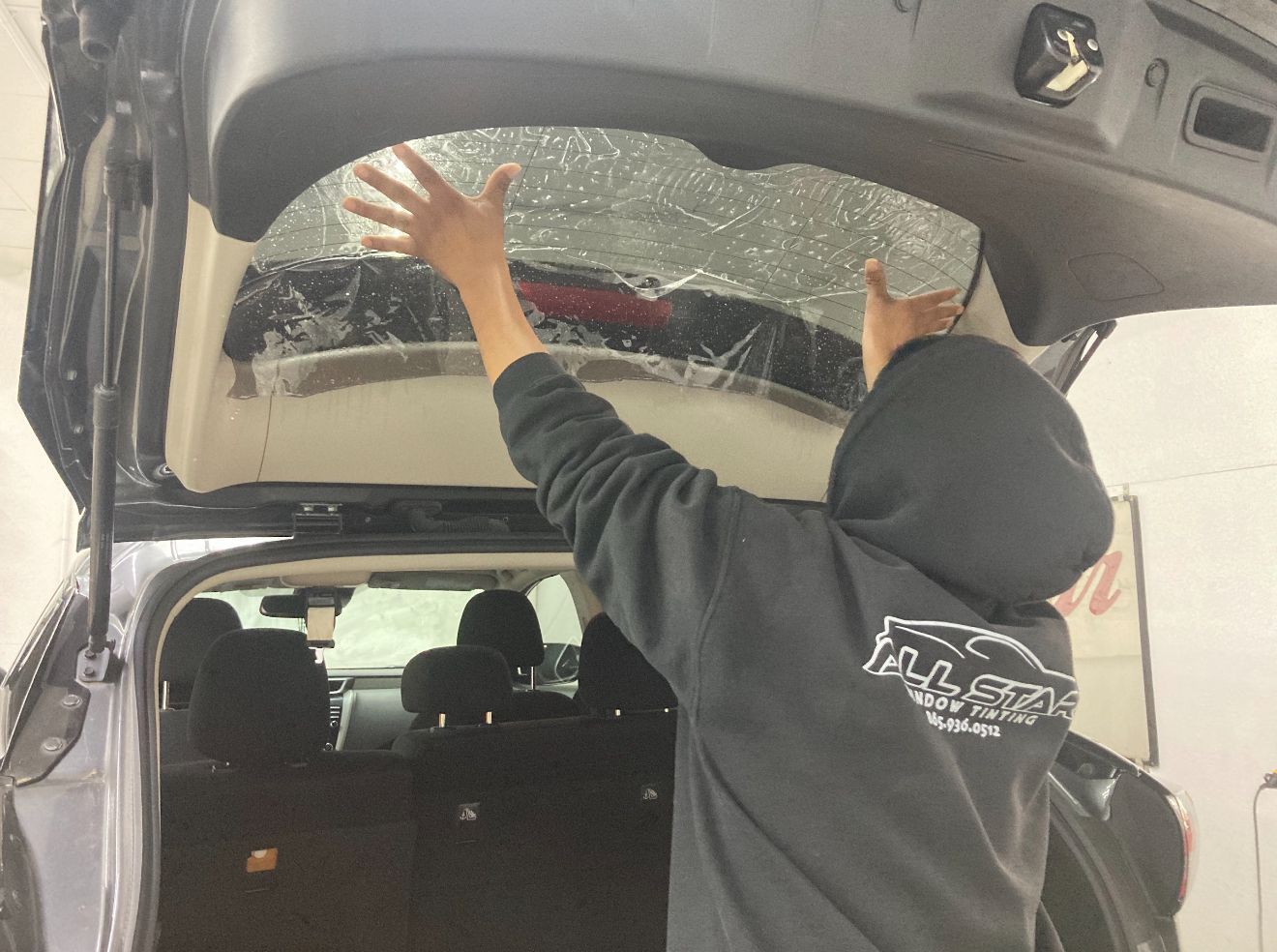 Person installing window tint on a car's rear window; arms raised, vehicle's trunk open.