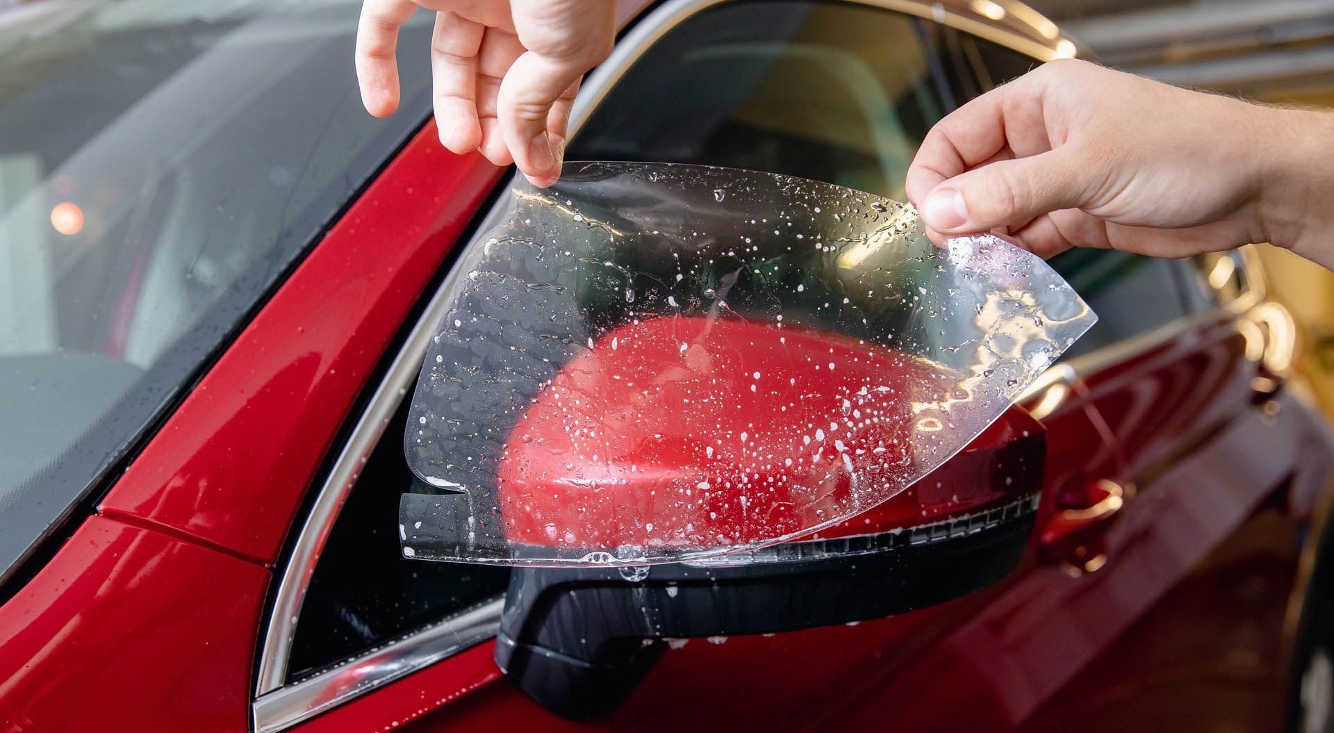 Hands peeling a clear protective film from a red car side mirror.