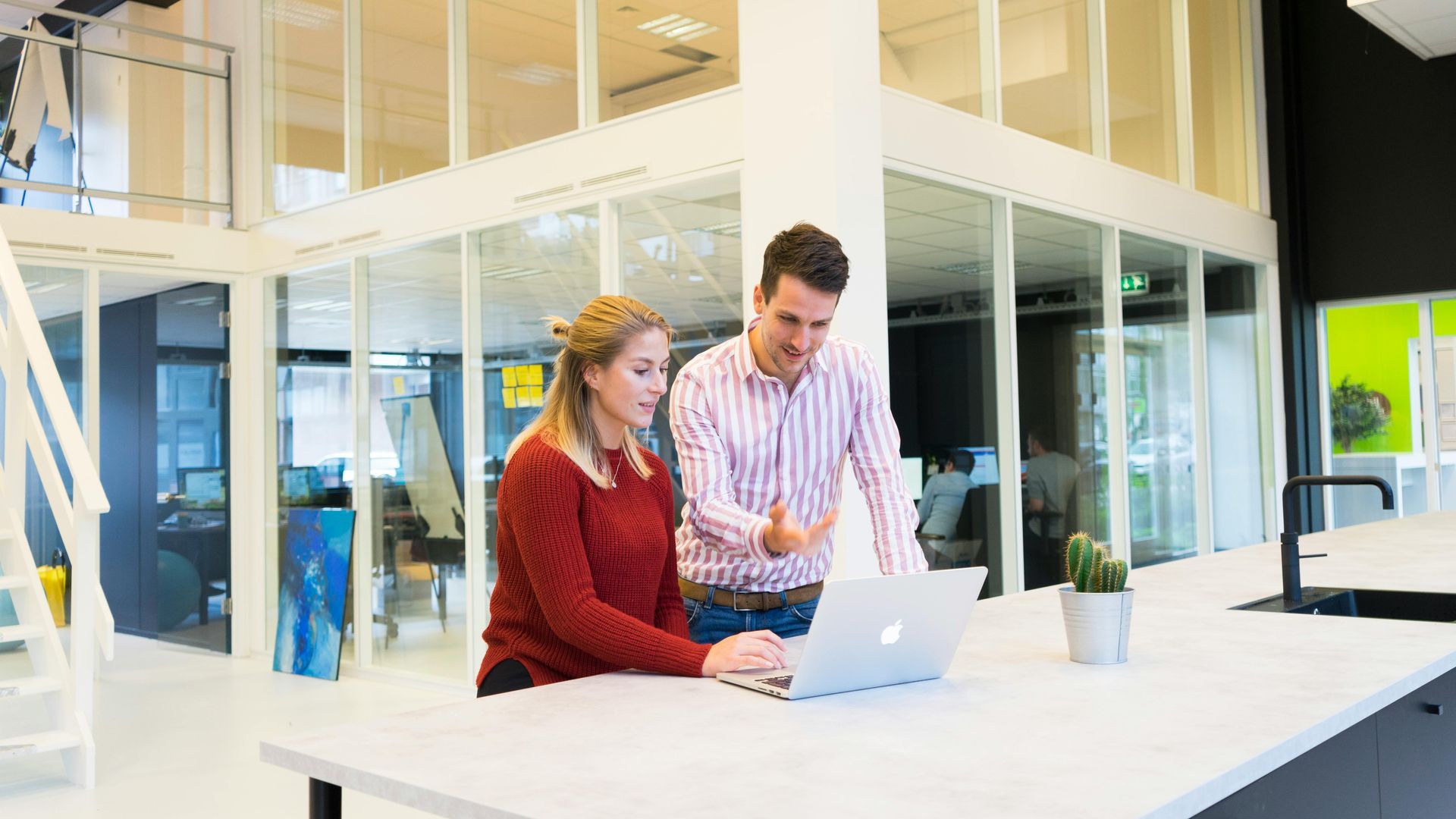 Woman and man look at a laptop in an office setting. The man gestures with his hand.