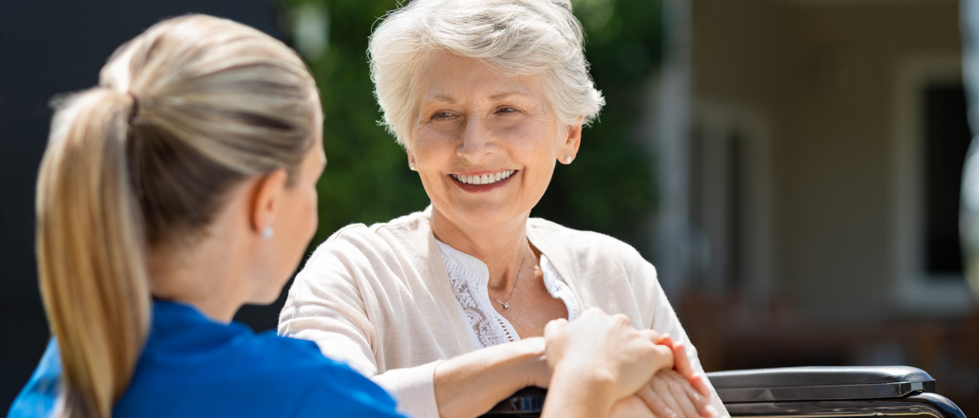 Uma cuidadora conforta uma senhora idosa sentada ao ar livre. A mulher sorri, com as mãos juntas em oração.
