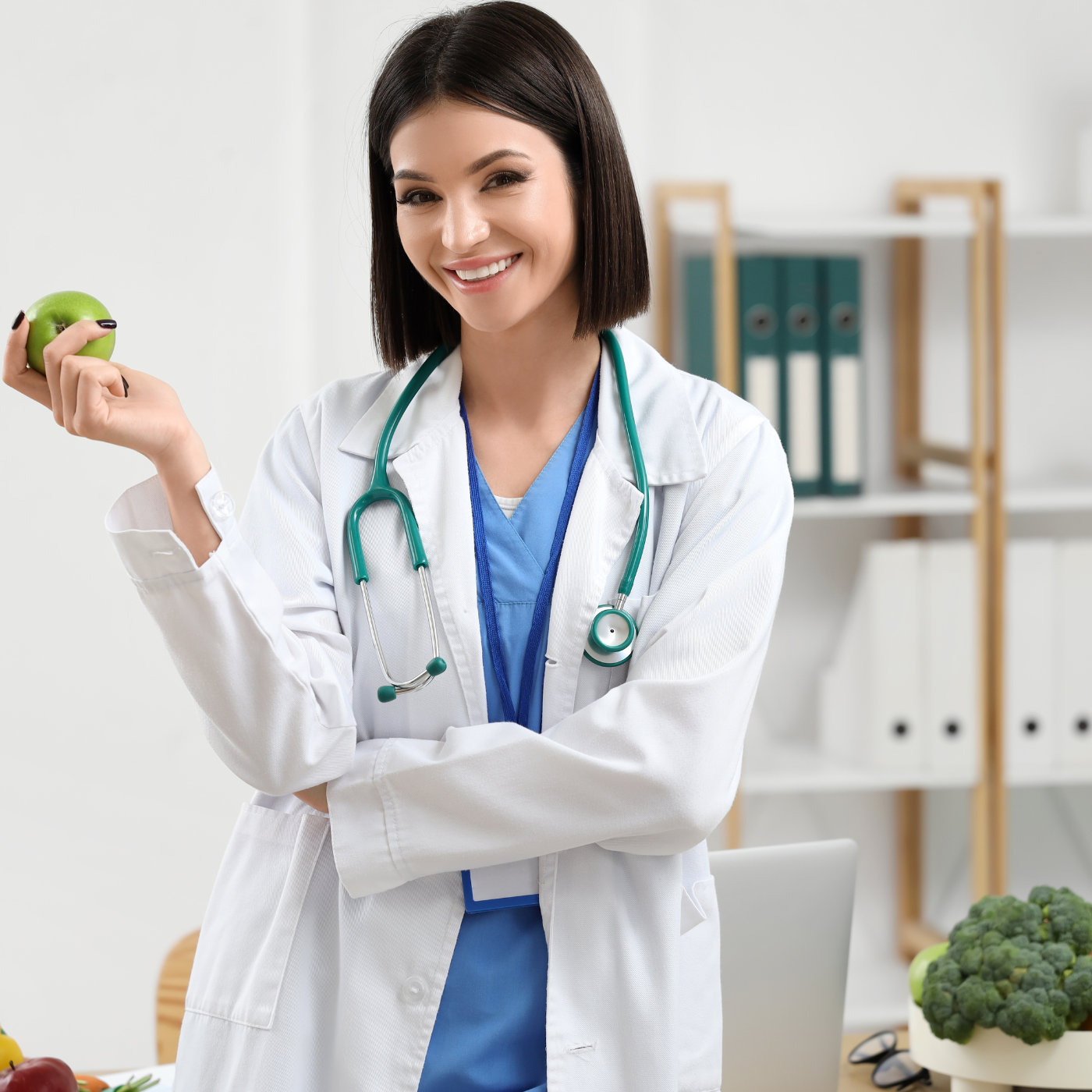 Uma nutricionista sorridente, de jaleco branco, segura uma maçã e um estetoscópio perto de uma mesa com frutas e brócolis.