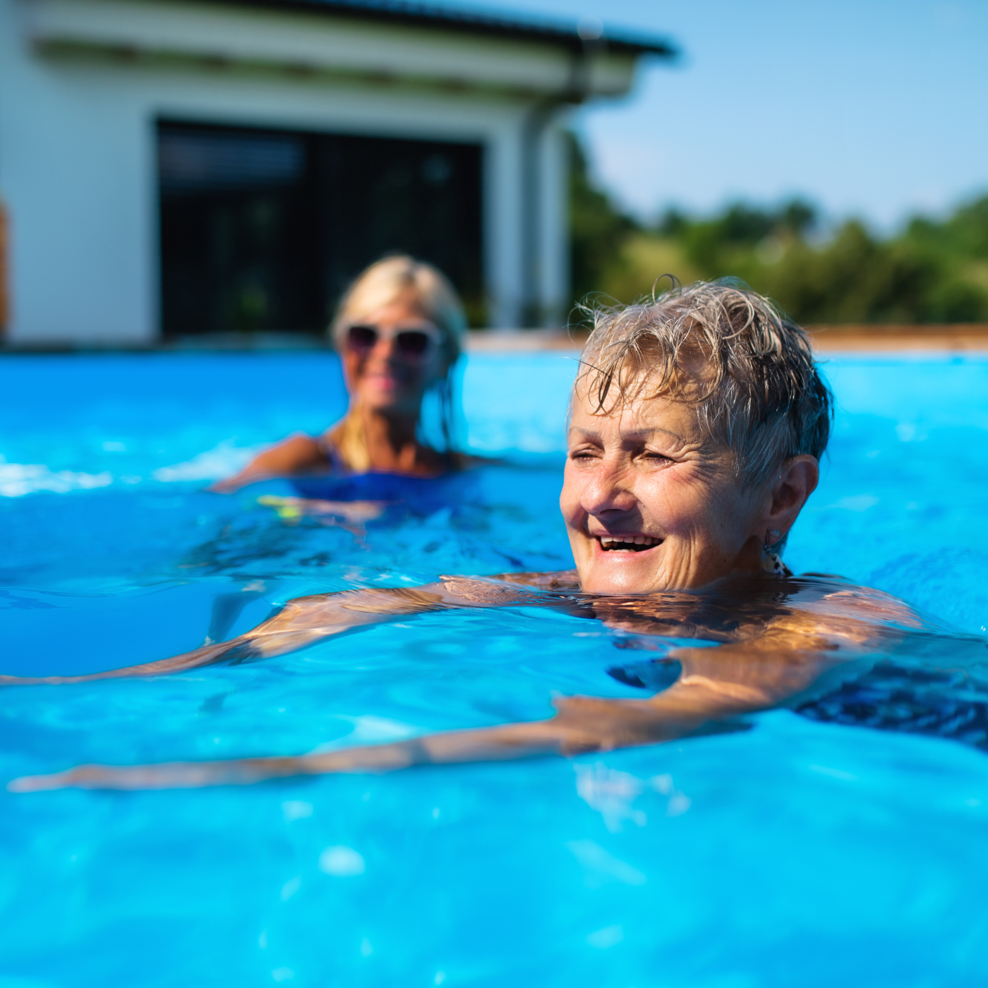 Duas mulheres sorrindo enquanto nadam em uma piscina azul, perto de uma casa em um dia ensolarado.