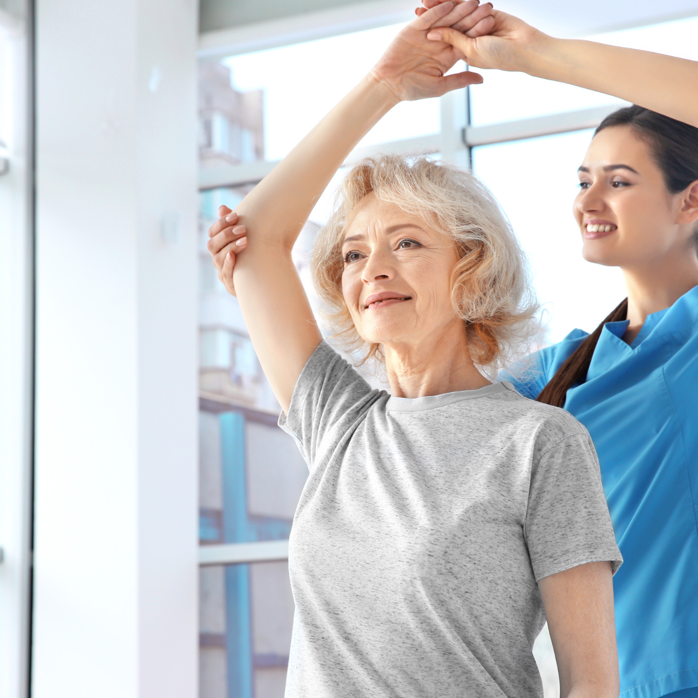 Mulher levanta o braço com a ajuda da terapeuta em sala bem iluminada, com janelas visíveis.