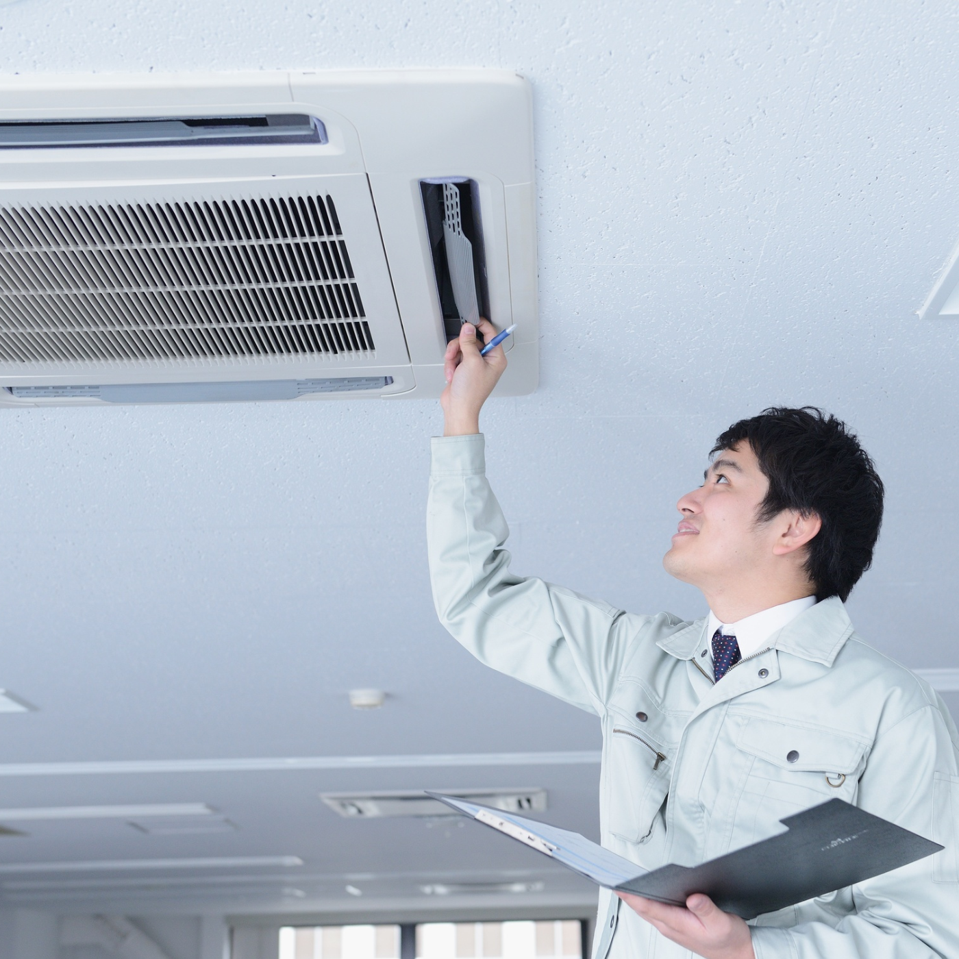 Homem com roupas de trabalho leves inspecionando um ar-condicionado de teto com uma prancheta em um ambiente de escritório.