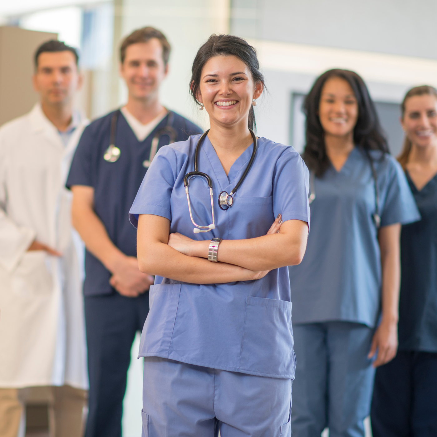 Equipe médica de jaleco branco e uniforme azul, sorrindo, braços cruzados. Ambiente interno e bem iluminado.