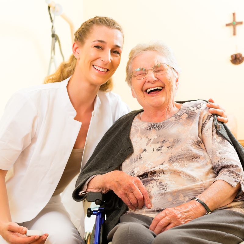 Uma mulher de uniforme médico sorri para uma pessoa idosa em cadeira de rodas. Ambas riem na sala.