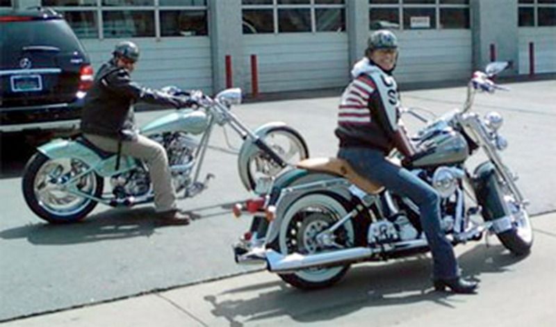 Two people wearing helmets sit on custom motorcycles parked in front of a building.