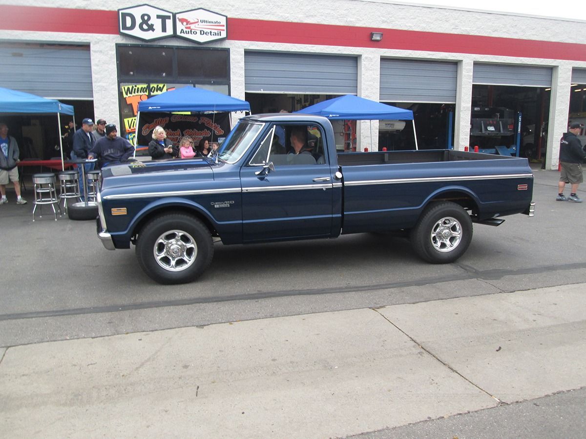 A dark blue vintage Chevrolet pickup truck parked outside an auto shop, with people visible in the background.