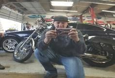 A person in a garage crouches while holding a smartphone up to take a photo of a motorcycle and car.