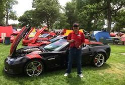 A person in a red shirt stands next to a black sports car with its hood open at an outdoor car show.