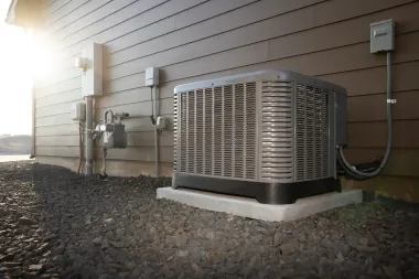Outdoor HVAC unit beside a house wall on a gravel bed in bright sunlight