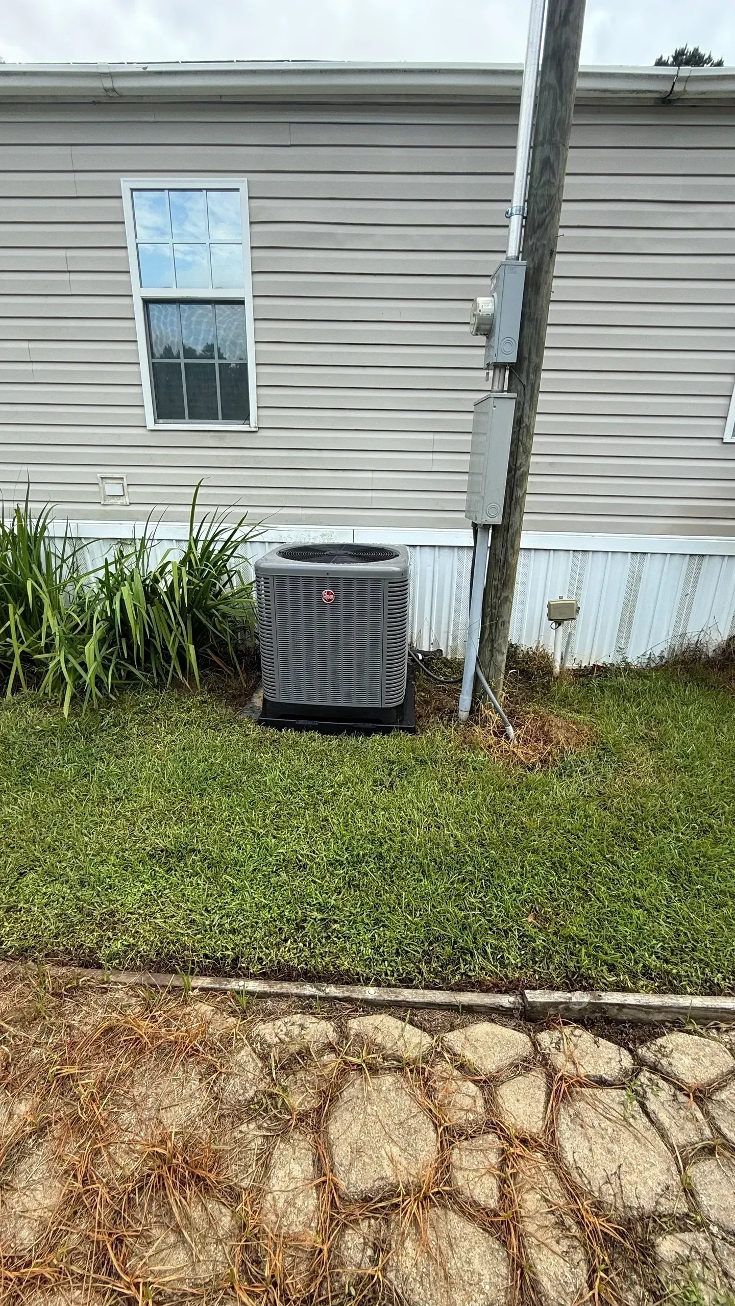 House exterior with beige siding, window, air conditioner unit, tree, and grassy yard with stone path