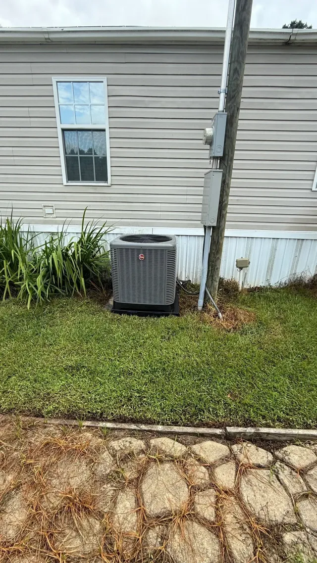 Outdoor air conditioner unit beside a tan brick wall on a grassy lawn, with utility meters nearby
