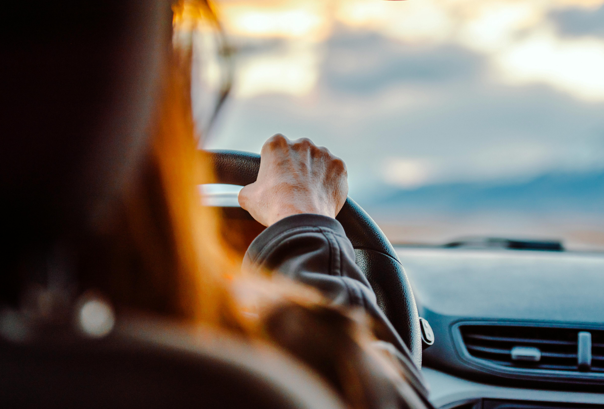 Person's hands grip a steering wheel, driving. Blurred background shows sky and mountains.