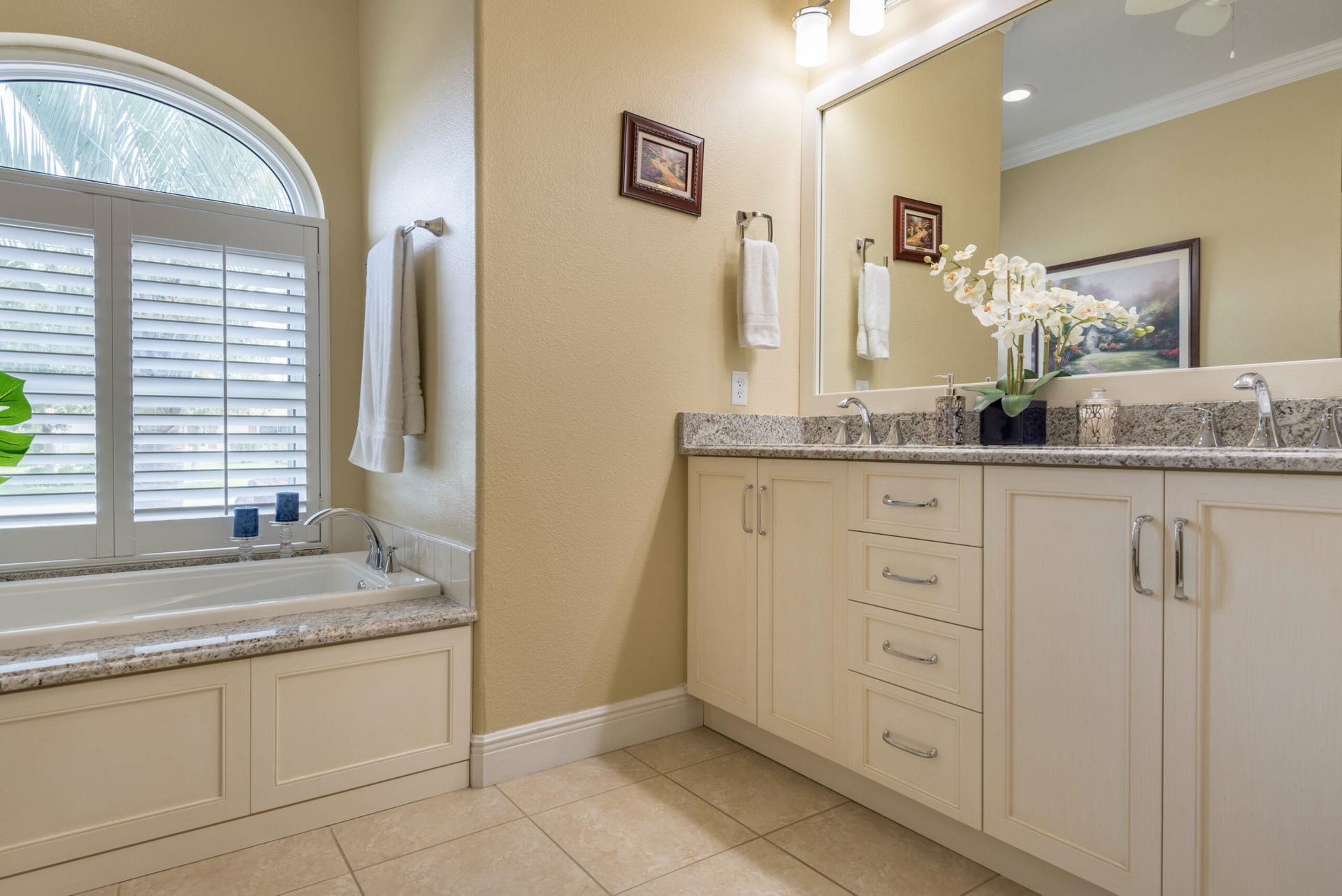 Bright beige bathroom with double vanity, large mirror, and arched window above a built-in tub