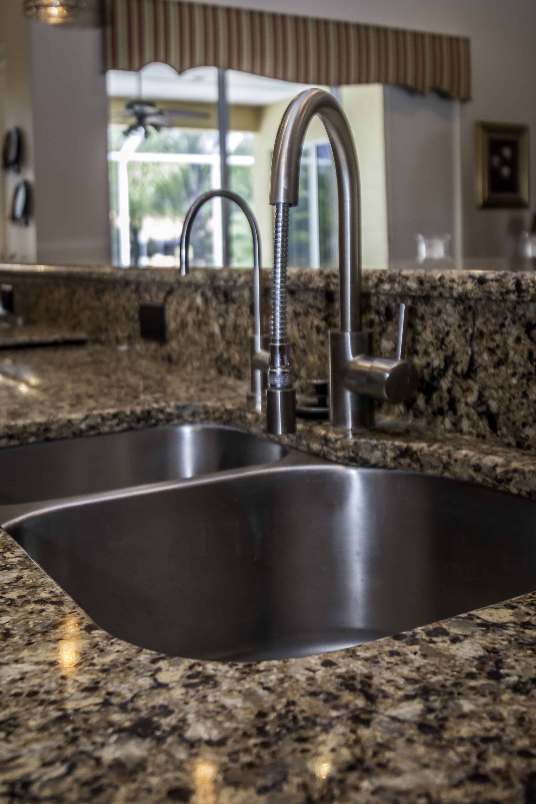 Granite kitchen sink with tall chrome faucet and a mirror reflecting a bright window