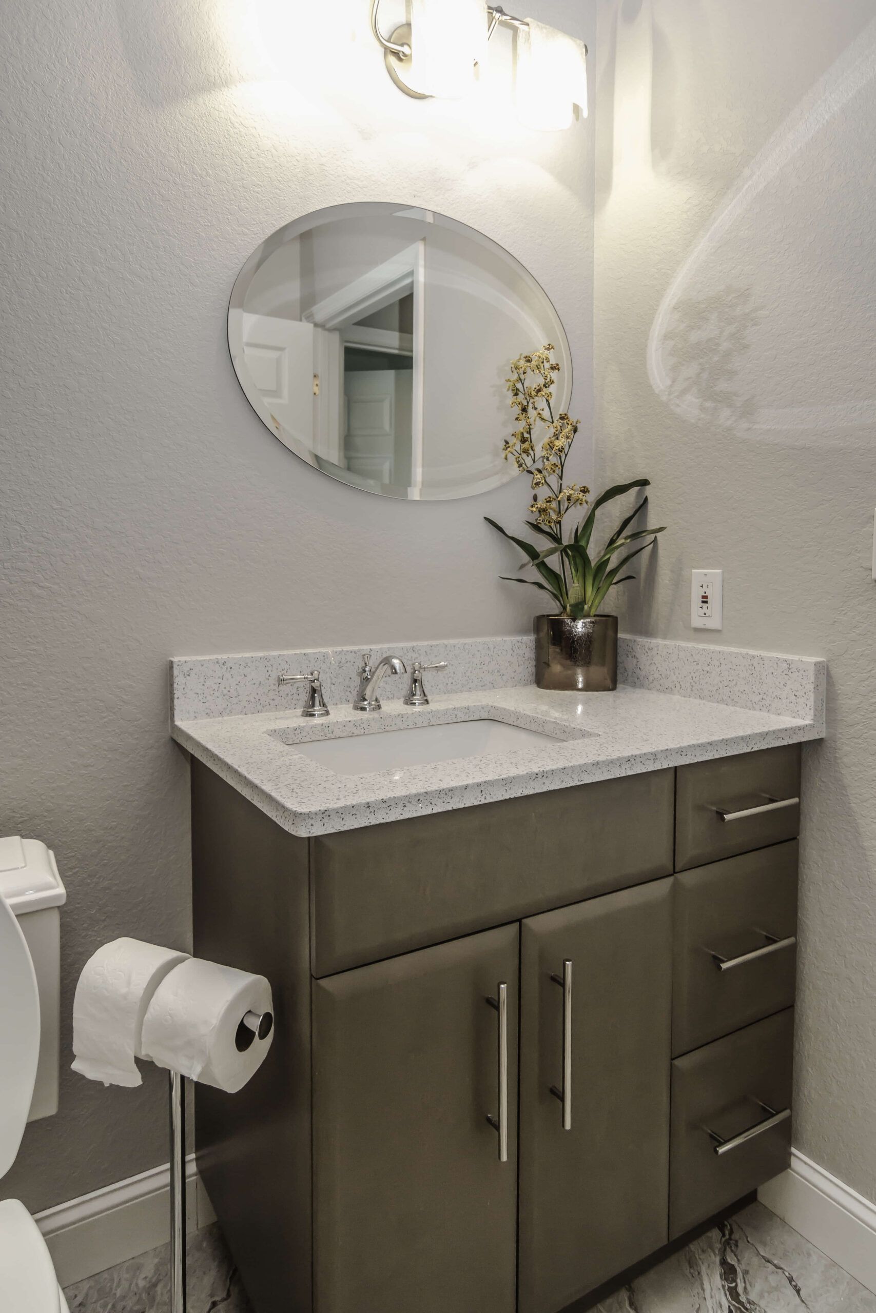 Small bathroom vanity with speckled countertop, round mirror, and flower arrangement beside a toilet paper holder.