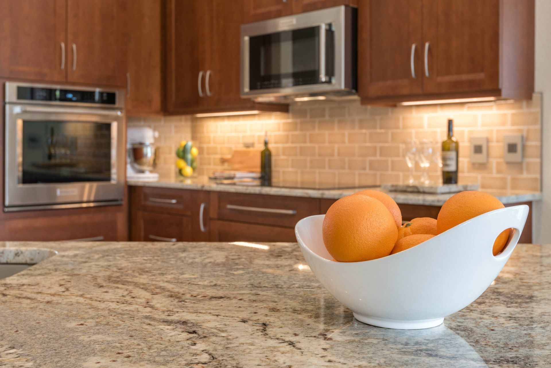 White bowl of oranges on a granite kitchen counter with wooden cabinets and stainless steel appliances in the background