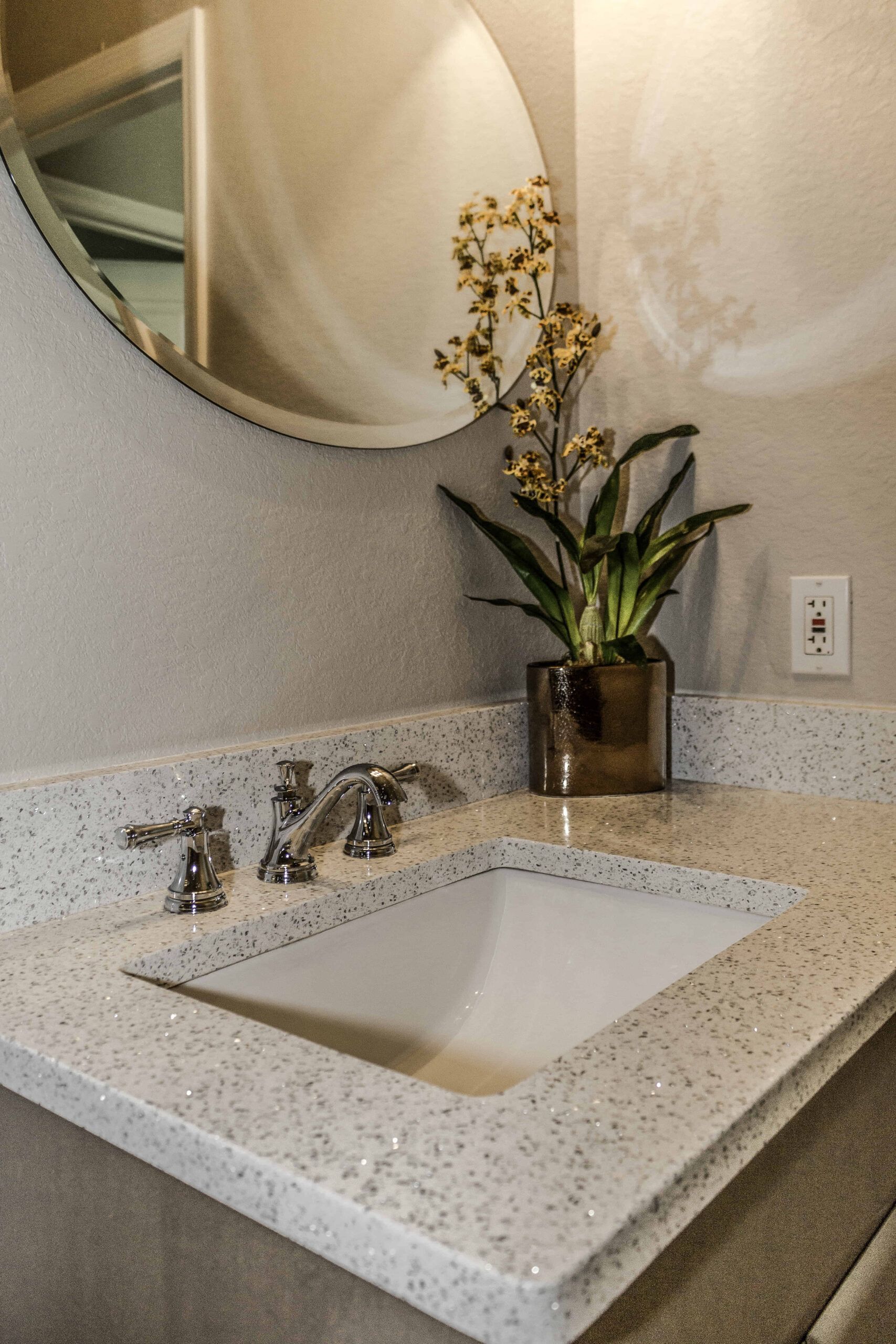 Small bathroom vanity with speckled countertop, white sink, chrome faucet, mirror, and orchid centerpiece
