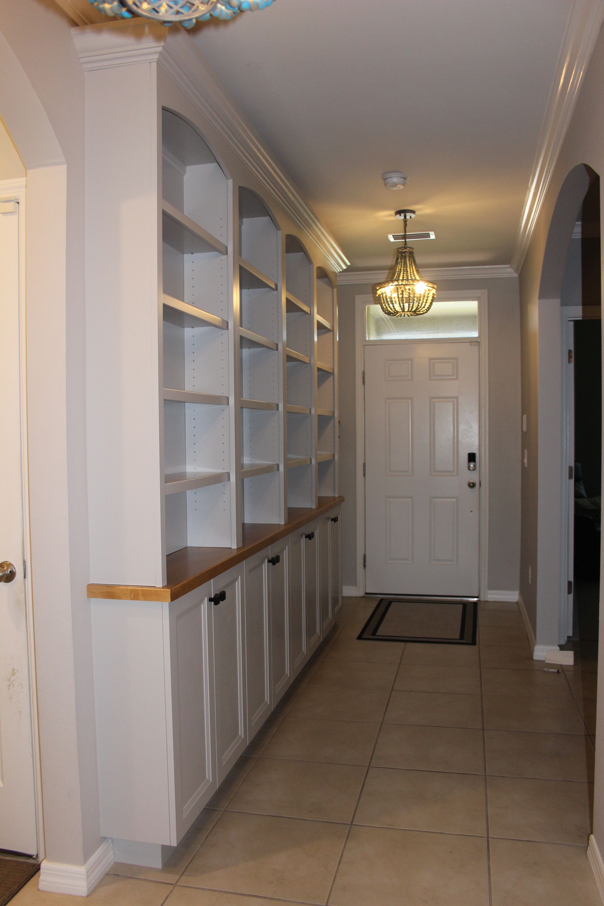 Long hallway with built-in white shelves, tiled floor, chandelier, and a closed white door at the end.