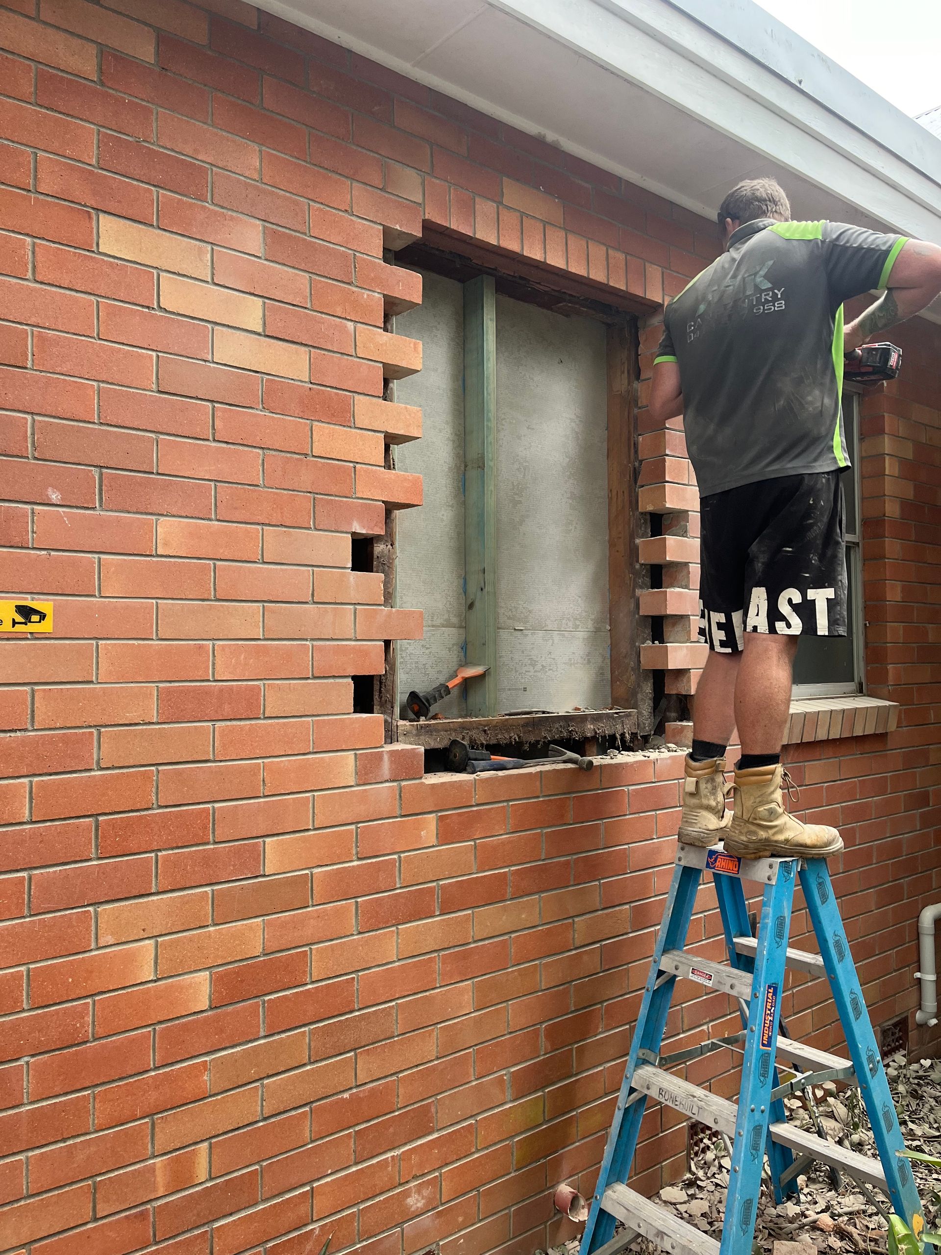 A man is standing on a ladder in front of a brick wall — TM Built Construction In Lochinvar, NSW