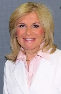 Woman with blonde hair, smiling, wearing a white jacket and pink shirt.